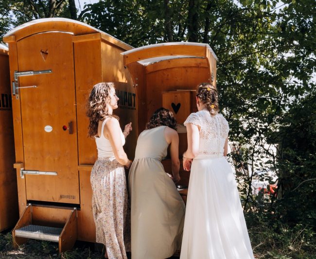 Kompotoi mit Waschbecker an dem gerade braut und Freundinnen sich die Hände waschen