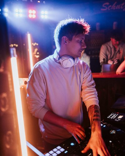 KILLI am DJ Pult mit Lichtröhren im Hintergrund
